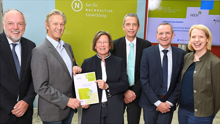 Foto mit sechs Personen, von denen zwei die Broschüre zum Hochschul-Nachhaltigkeitskodex in die Kamera halten und vor Bilderwand des Rats für nachhaltige Entwicklung sowie HOCH-N stehen.